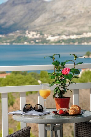 A table with a book, glasses, a glass of orange juice, a potted plant, and pastries on a balcony overlooking a body of water and mountains. - Villa Nick-sunrise Deluxe Apartment (Konavle)