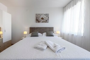 A neatly made bed with two towels, a headboard, bedside lamps, and a floral artwork on the wall. - Villa Nick-sunrise Deluxe Apartment (Konavle)