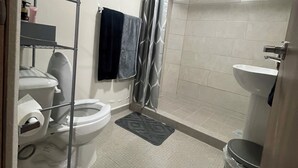 Un baño con inodoro, ducha, lavamanos y una estantería metálica.