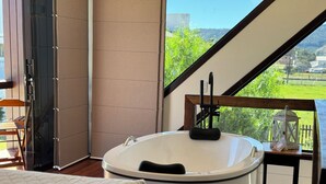 A modern bathroom with a freestanding bathtub, a wooden chair, and a view of greenery outside.