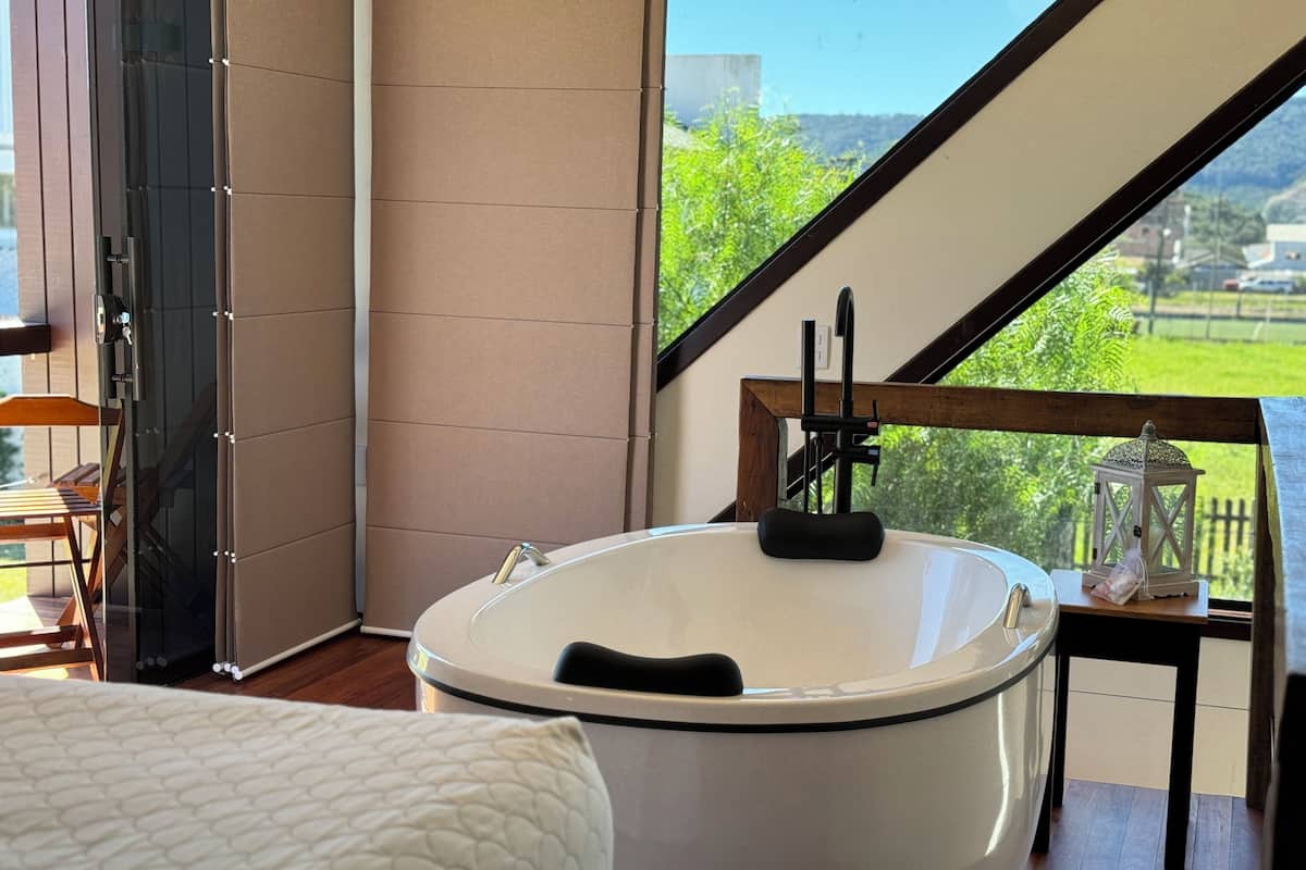 A modern bathroom with a freestanding bathtub, a wooden chair, and a view of greenery outside.