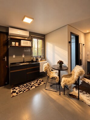 A modern kitchen with a black countertop, a microwave, and a window with blinds. There is a round table with two chairs and a small rug on the floor. - Lofts Urbano (Urubici)