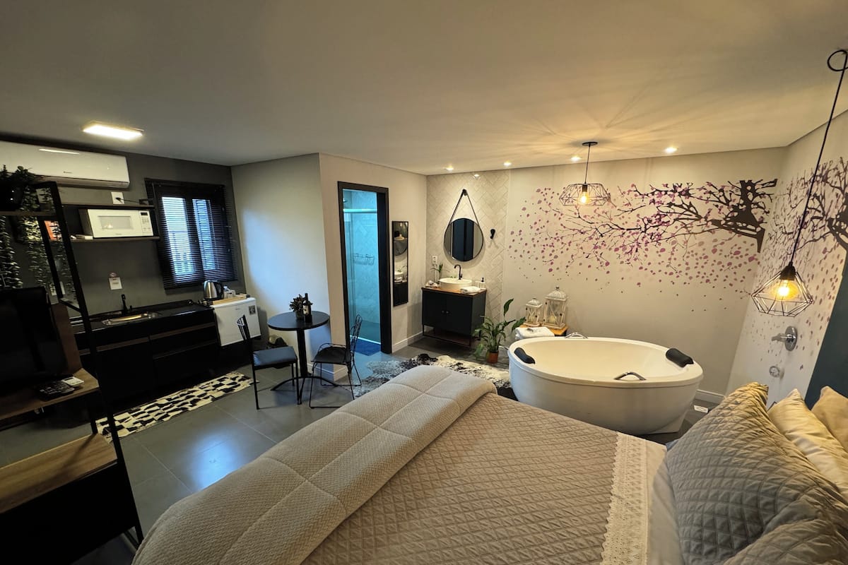 A modern bedroom with a large bed, a separate bathroom, a small dining area, and a kitchenette.