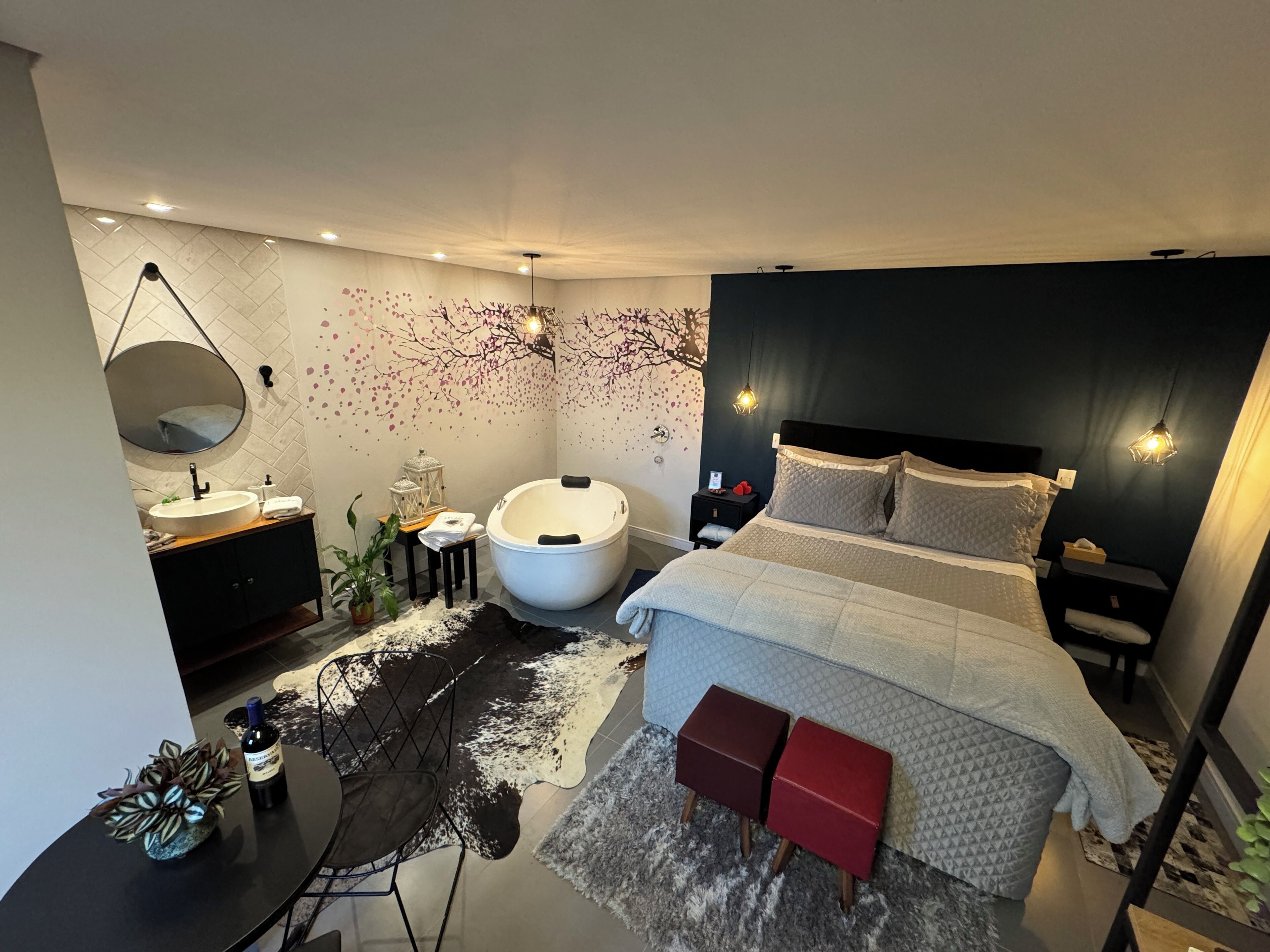 A modern bedroom with a large bed, a round bathtub, a vanity with a mirror, and a small stool.
