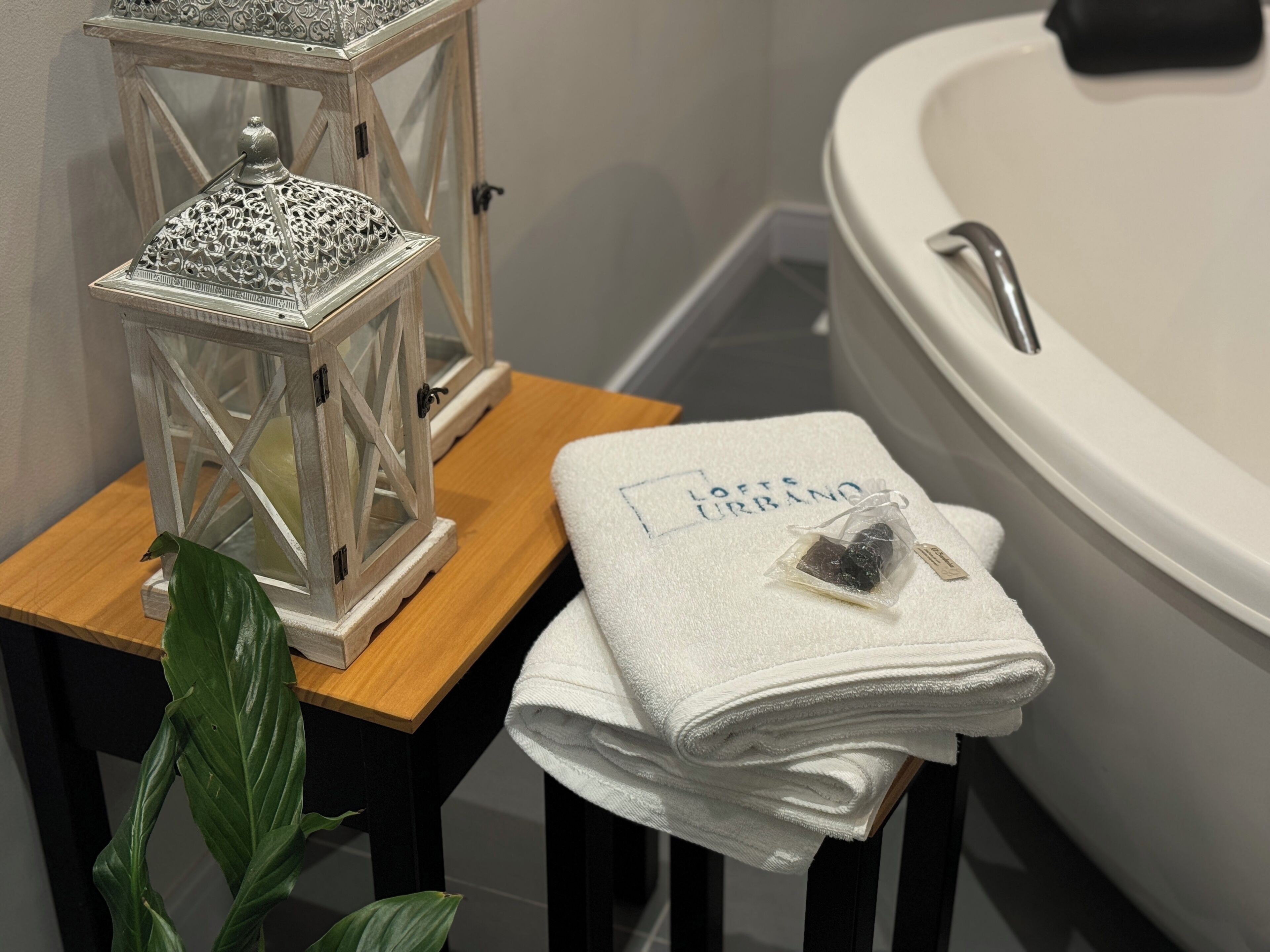 Un baño con una bañera blanca, un taburete de madera y una toalla blanca doblada, junto con un farol decorativo.