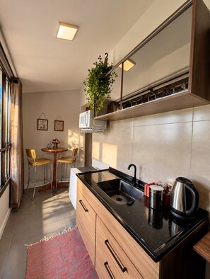 A compact kitchen with a black countertop, a stainless steel kettle, and a microwave. There is a small dining area with two chairs and a table. - Lofts Urbano (Urubici)
