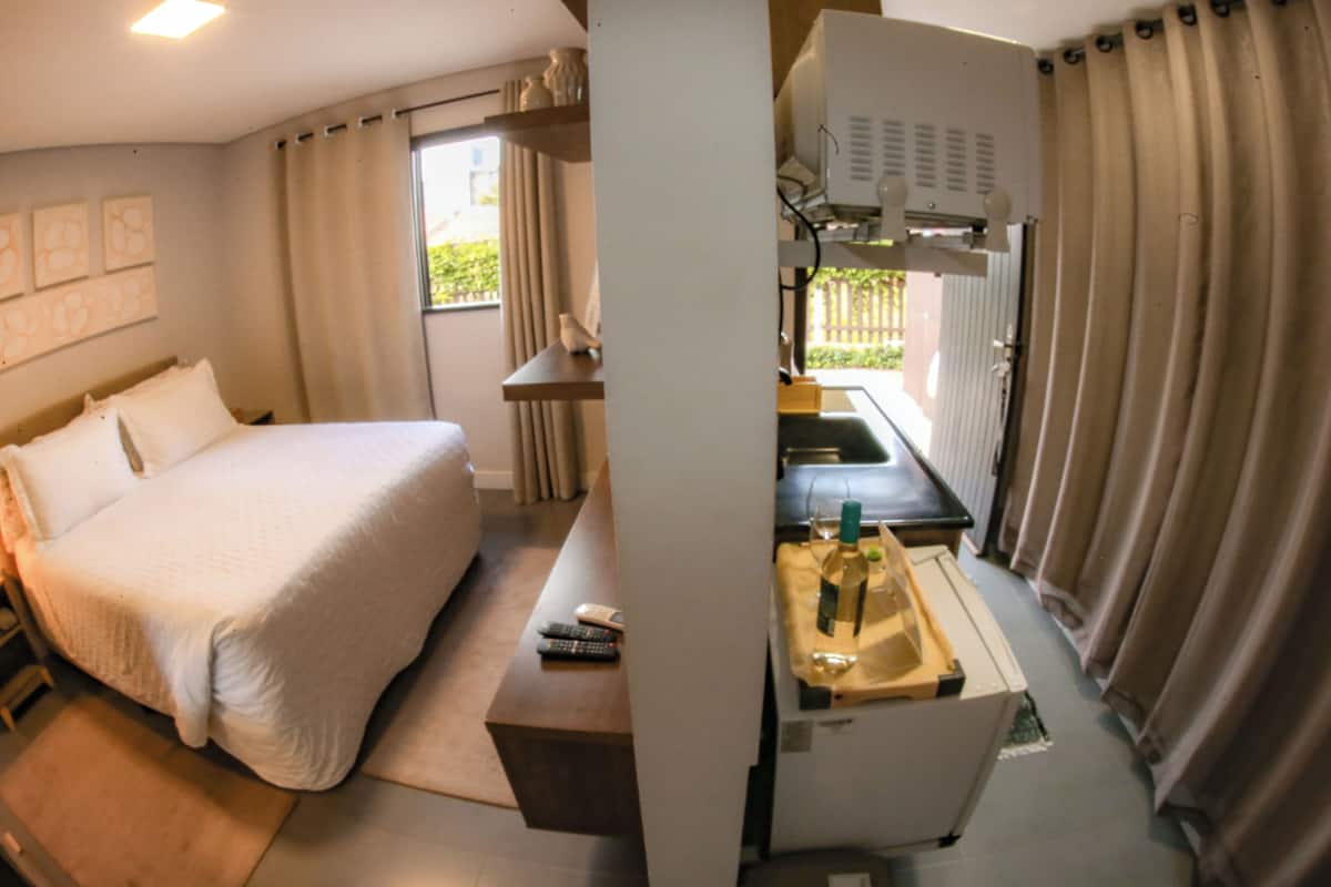 A hotel room with a bed, a desk, a chair, a small table with a bottle and glass, and a window with curtains.