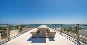 Un banco de concreto con barandilla de vidrio que da a una playa con palmeras y el océano.