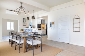 A dining area with a round table, chairs, and pendant lighting. There is a kitchen in the background with white cabinets and a hanging rack. - Luxury Beachfront Home: Pool, Large Groups, Boat, Water Sports (San Pedro)