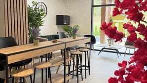 Un espace repas moderne avec une table en bois, des chaises noires et des tabourets. On y trouve également une plante en pot et un téléviseur fixé au mur.
