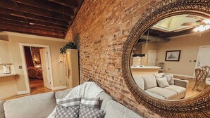 Un salón con pared de ladrillo, un espejo grande, un sofá con cojines estampados y una puerta que da a otra habitación.