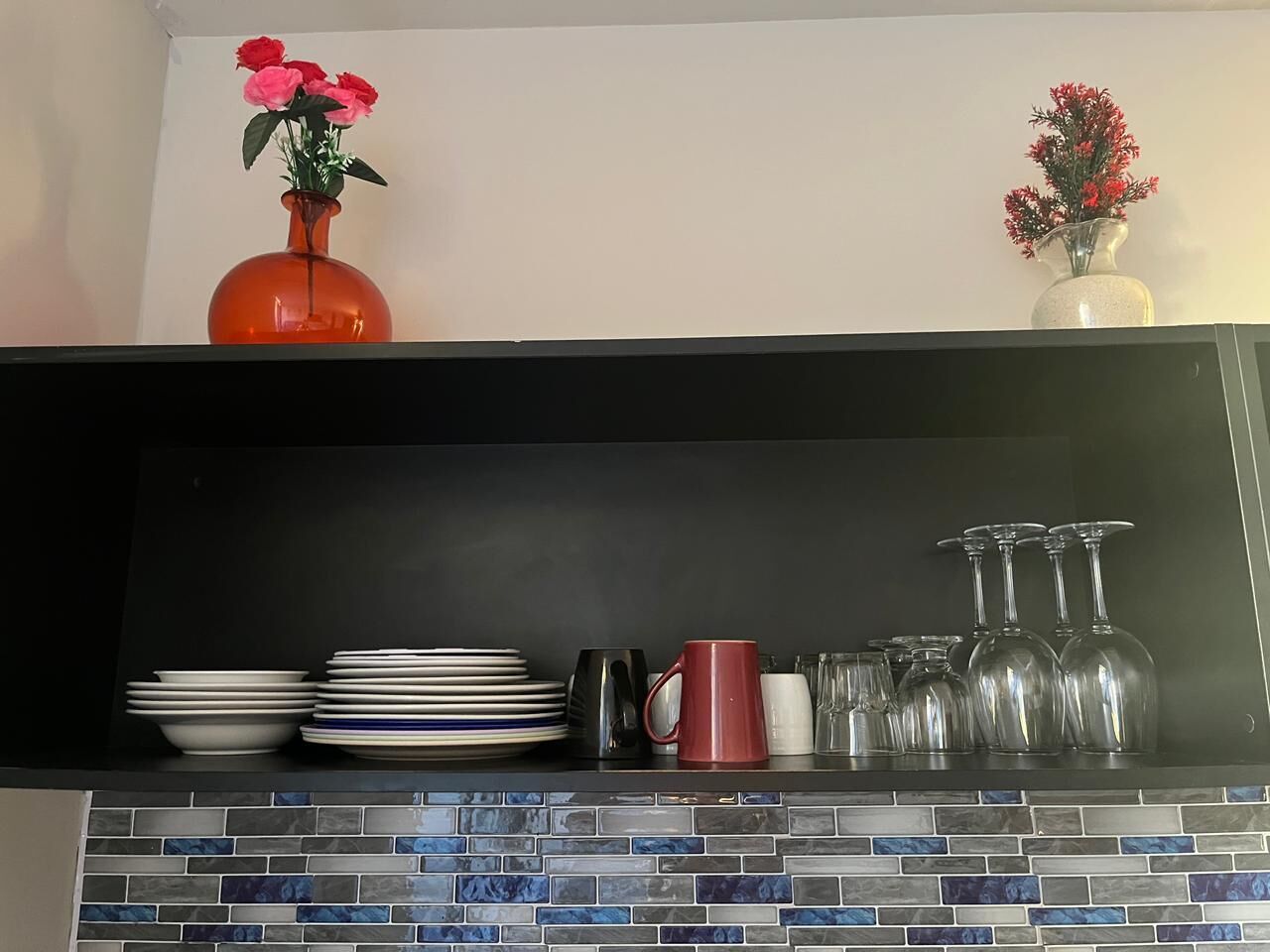 A kitchen counter with a tiled backsplash, a black shelf, and various kitchenware including plates, a red mug, and glassware.