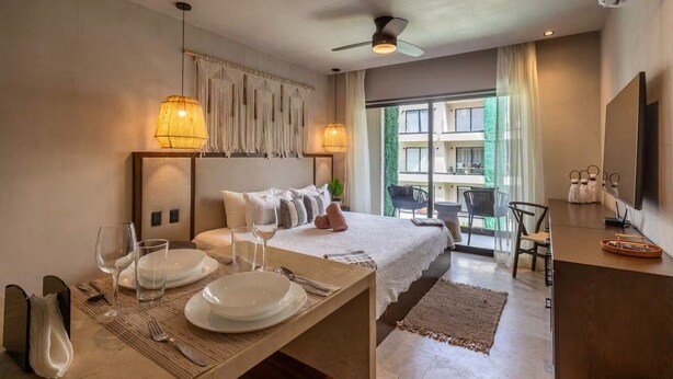 A hotel room with a bed, a dining table set for two, a TV, and a balcony with a chair. - Deluxe studio in downtown Tulum with pool, rooftop and gymnasium (Tulum)
