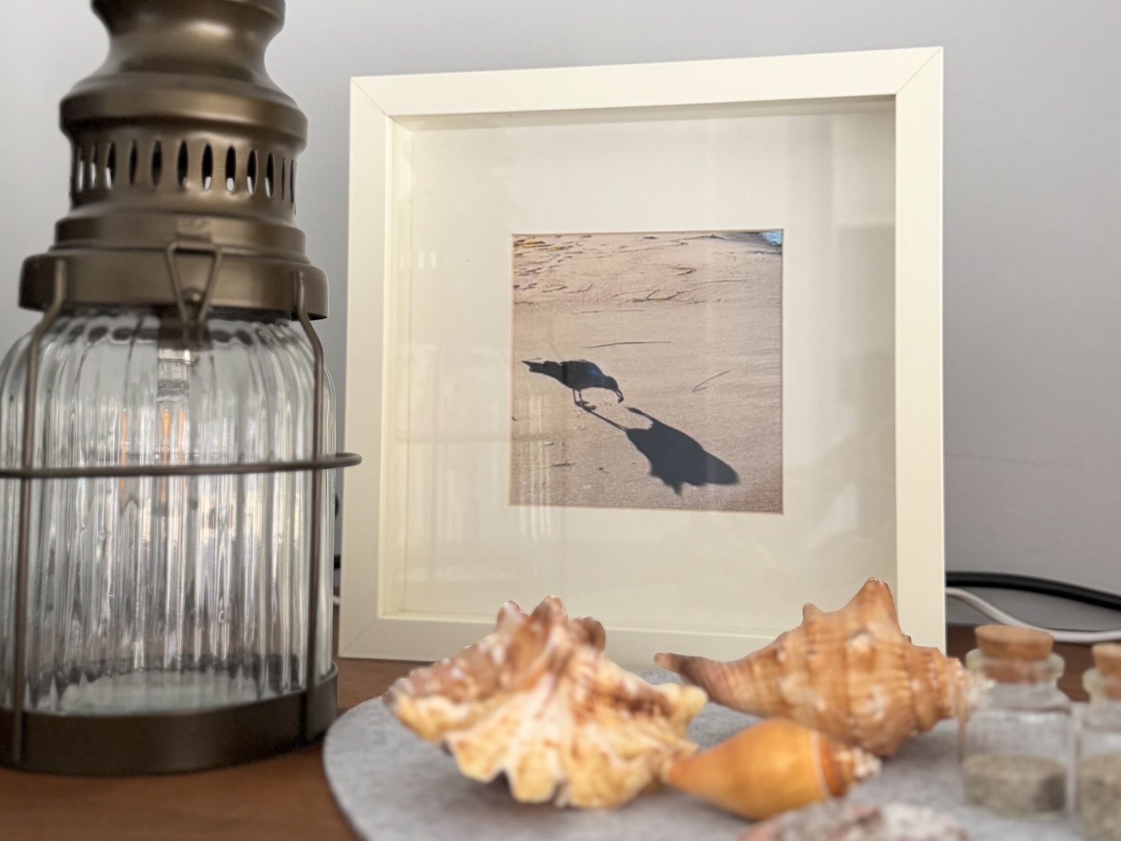 A framed artwork of a bird on a beach, a glass lantern, and seashells on a table.