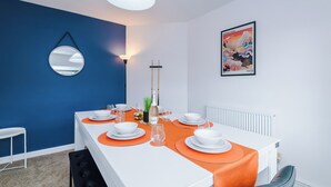Ein zeitgemäßes Esszimmer mit einem weißen Tisch, gedeckt für vier Personen, einer blauen Akzentwand, einem hängenden Spiegel und einem gerahmten Kunstwerk.