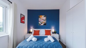 Ein Hotelzimmer mit einem Bett, weißen Nachttischen, roten und weißen Kissen, einer blauen gesteppten Bettdecke, einem Bild an der Wand und einem Fenster mit weißen Vorhängen.