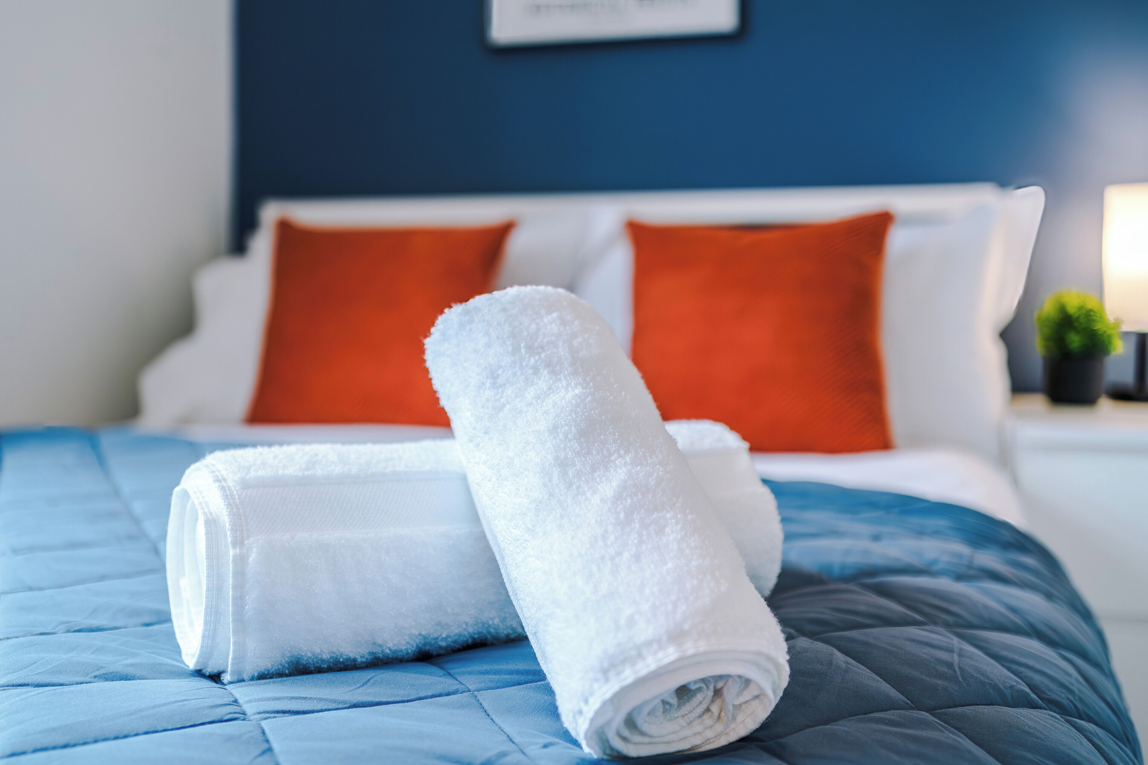 Una cama bien hecha con un edredón azul, dos toallas blancas y dos almohadas naranjas.