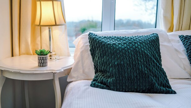 A neatly made bed with a green knitted pillow, a bedside table with a lamp, and a small potted plant. - Snug 3BR House | Contractors | Parking | Pool Table (Rolleston-on-Dove)