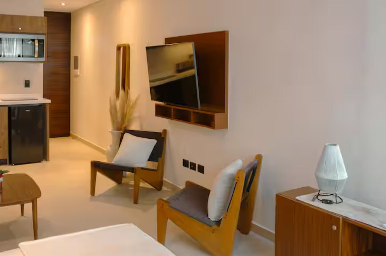 Un salon moderne avec un téléviseur à écran plat fixé au mur, une table basse en bois, deux fauteuils, un petit meuble et une lampe.