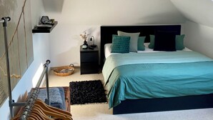 Ein Schlafzimmer mit schrägen Wänden, einem Bett mit türkisfarbenen Bettwäsche, einem Nachttisch mit Lampe und einem Holzgestell mit Kleiderhaken.