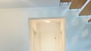 Ein schmaler Flur mit einer weißen Tür am Ende, eine weiße Kommode mit einem schwarzen Gegenstand darauf und ein flauschiger weißer Sessel.