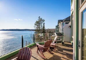 A patio with Adirondack chairs overlooking a lake, a grill, and a glass railing.