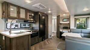 Une cuisine compacte intégrée au camping-car, comprenant une cuisinière, un four à micro-ondes et un évier, un coin repas avec une table et un banc, ainsi qu’une petite kitchenette équipée d’un réfrigérateur et d’un four.