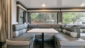 Ein kompaktes Wohnmobil-Interieur mit einem zentralen Tisch, gepolsterten Sitzgelegenheiten und großen Fenstern, die einen Blick auf Bäume bieten.