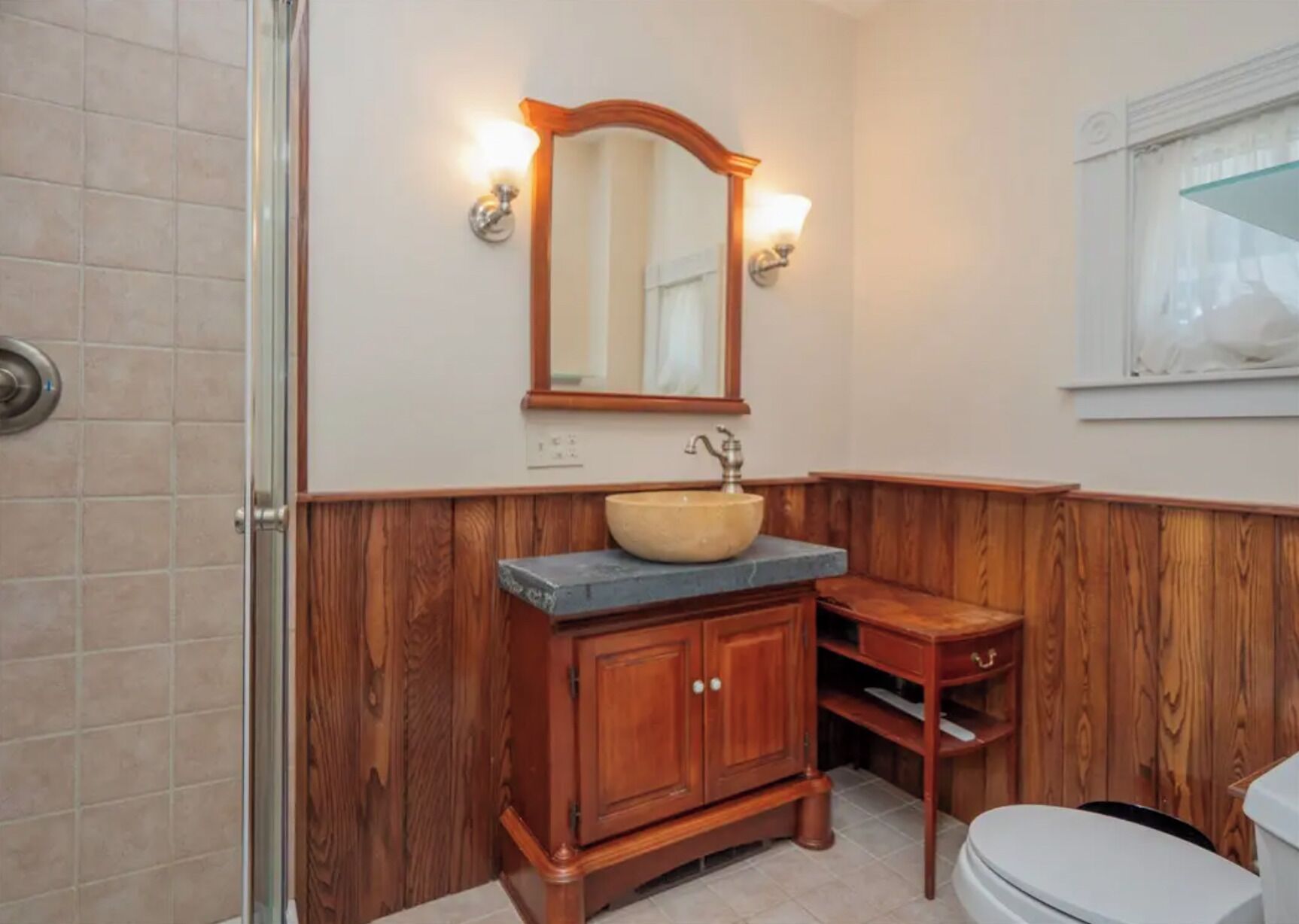 A bathroom with a wooden vanity, a vessel sink, a mirror with wall sconces, and a toilet.