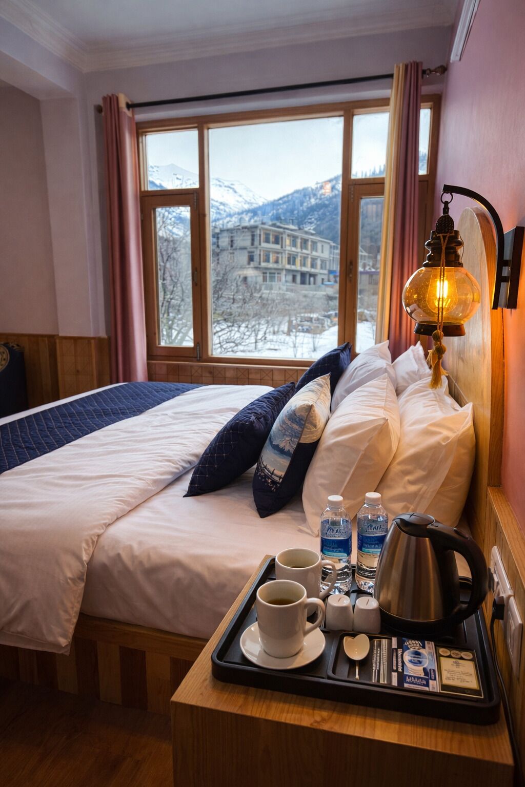 Une chambre d’hôtel avec un lit, une fenêtre donnant sur les montagnes, une table de chevet sur laquelle se trouve un plateau avec des articles pour le petit-déjeuner, et une lampe fixée au mur.