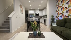 A modern living room with a white coffee table, a green sofa, and a dining area with a dark wooden table. - Furnished 3BR/3BA Brand New Anaheim Condo Move-In Ready & Utilities Included (Anaheim)
