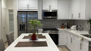 A modern kitchen with white cabinets, stainless steel appliances, and a central island with a plant. - Furnished 3BR/3BA Brand New Anaheim Condo Move-In Ready & Utilities Included (Anaheim)