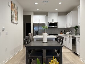 A modern kitchen with white cabinets, stainless steel appliances, and a dining area with a dark table and chairs. - Furnished 3BR/3BA Brand New Anaheim Condo Move-In Ready & Utilities Included (Anaheim)