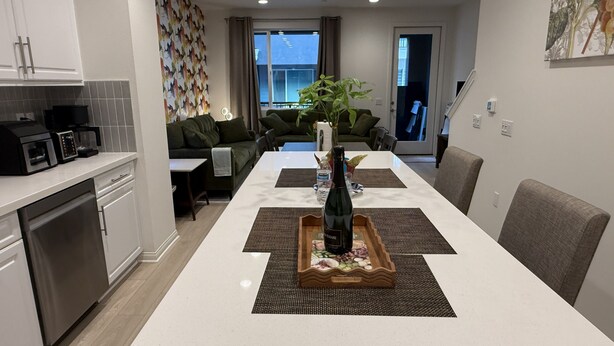 A modern kitchen with built-in appliances, a dining area with a long table, and a living room with a sofa and a plant. - Furnished 3BR/3BA Brand New Anaheim Condo Move-In Ready & Utilities Included (Anaheim)