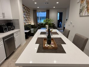 A modern kitchen with built-in appliances, a dining area with a long table, and a living room with a sofa and a plant. - Furnished 3BR/3BA Brand New Anaheim Condo Move-In Ready & Utilities Included (Anaheim)