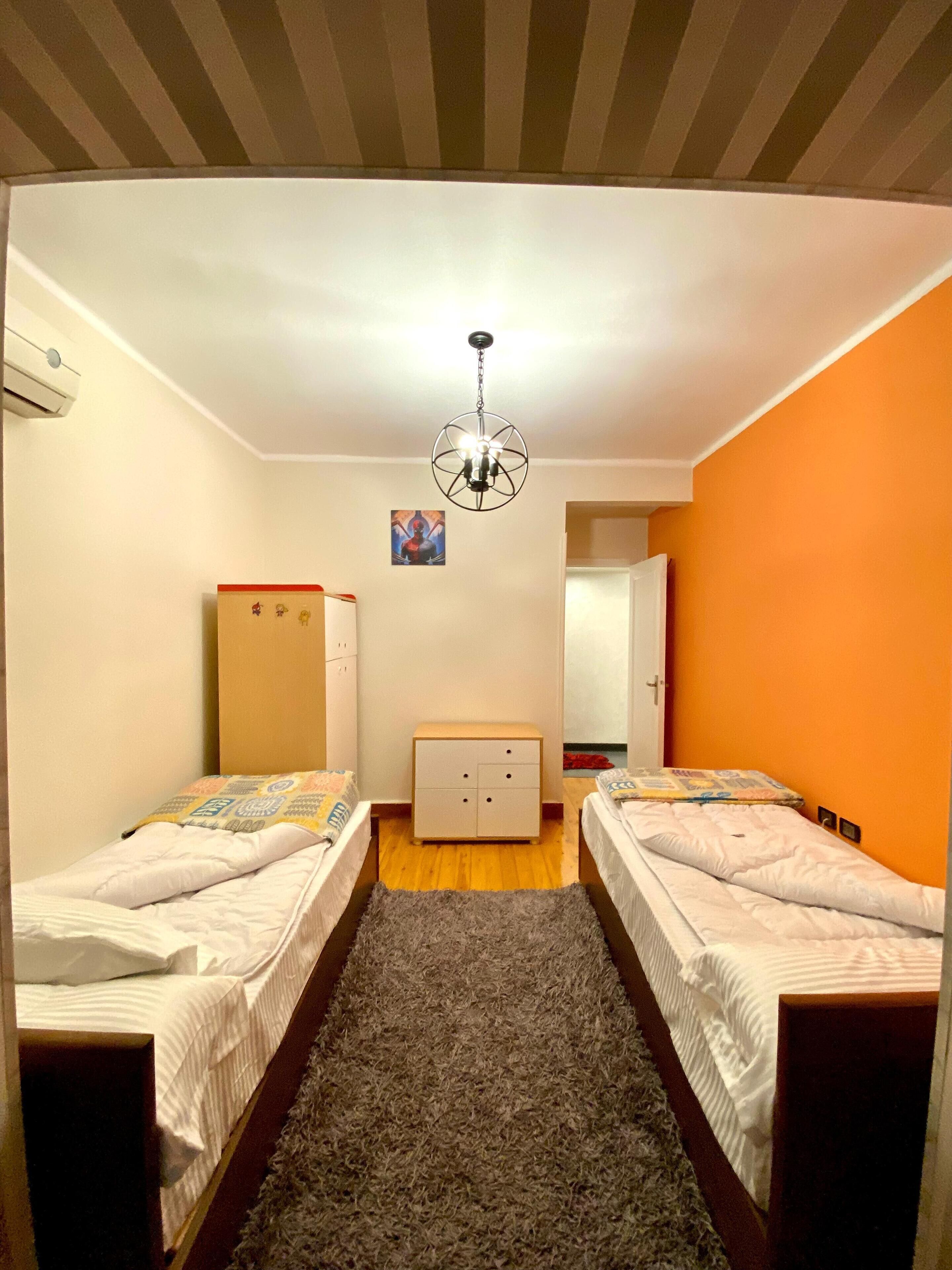 A room with two beds, a dresser, a chandelier, and a door leading to another room.
