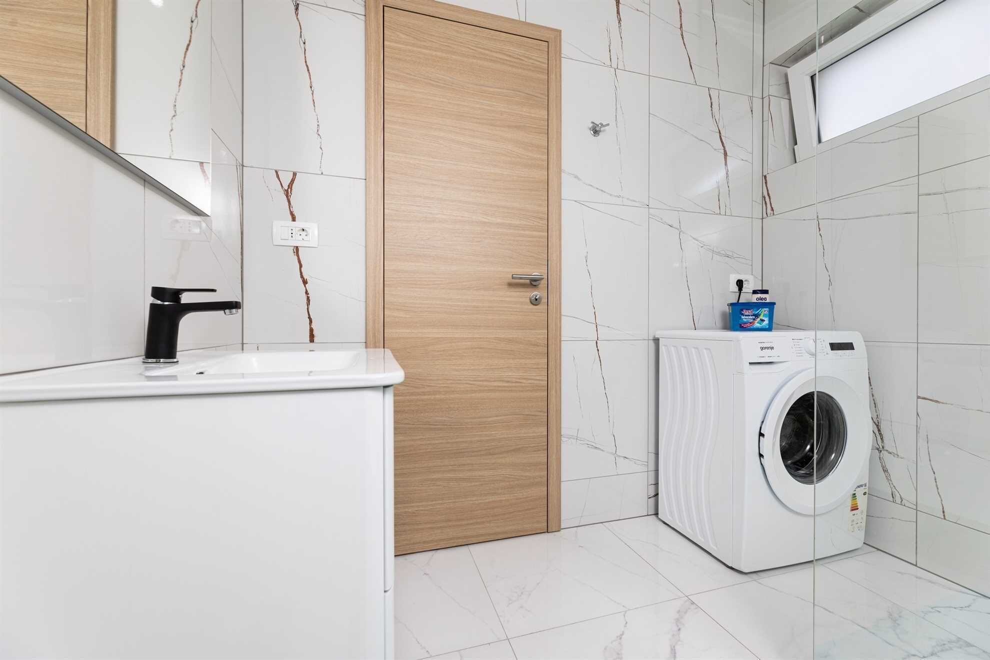 A modern bathroom with a sink, wooden door, white tiled walls, and a washing machine.