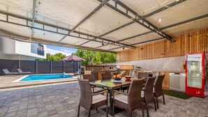 Un patio exterior con piscina, mesa de comedor, sillas y refrigerador.