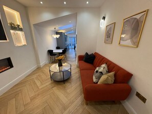 A modern living room with a herringbone floor, a red sofa, a glass coffee table, and a fireplace. - Stylish Kentish Town Home/Private Garden & Jacuzzi (London)