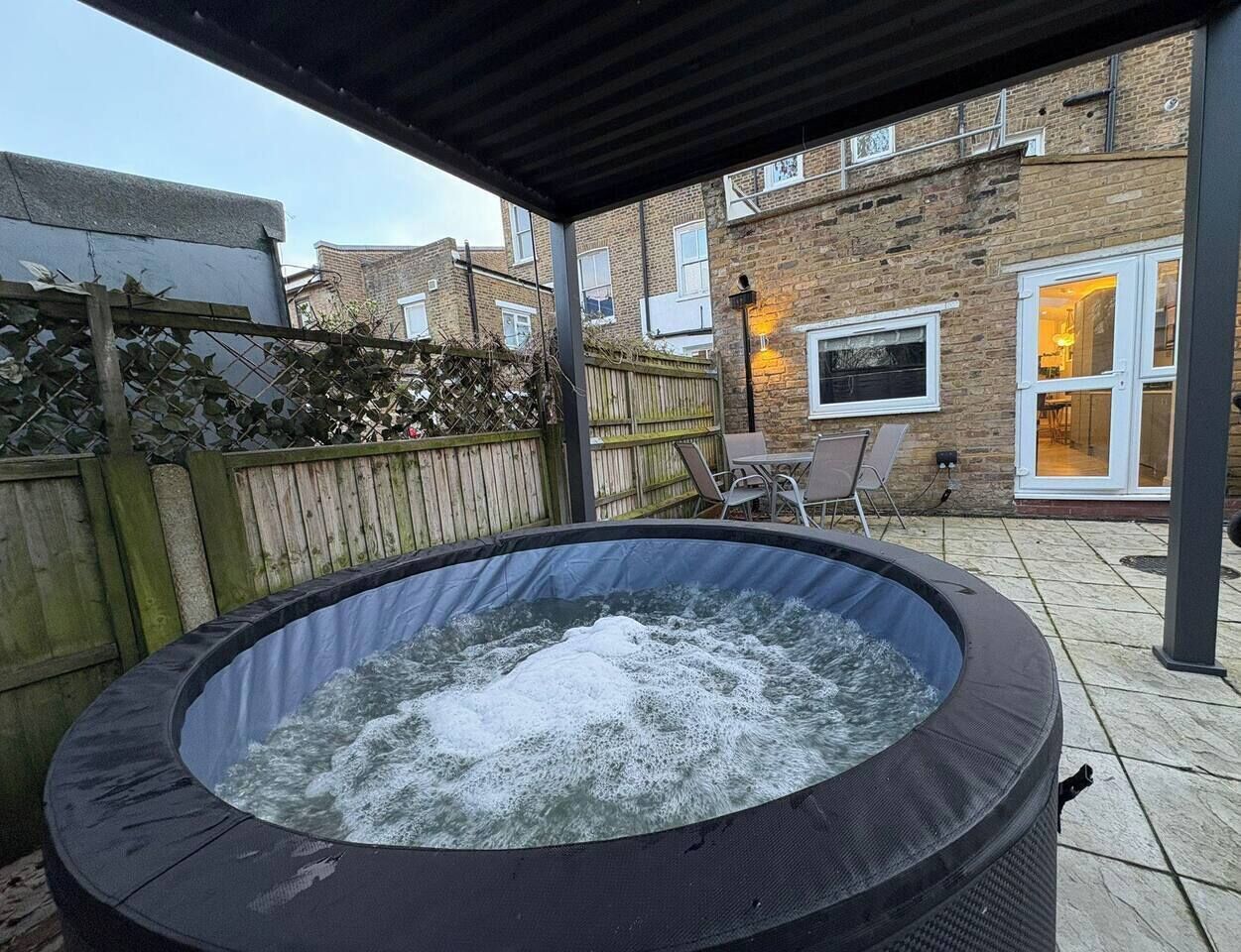 Un jacuzzi dans un jardin, avec une clôture en bois, un bâtiment en briques et une table entourée de chaises.