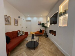 Ein modernes Wohnzimmer mit einem roten Sofa, einem orangefarbenen Sessel, einem Glastisch mit Blumen und einem Kamin.