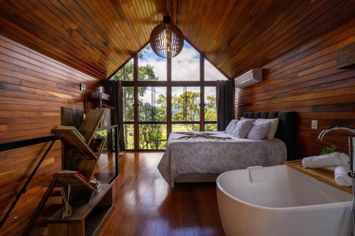 A wooden cabin bedroom with a bed, a bathtub, and a large window offering a view of the outdoors.