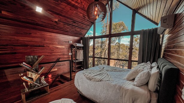 Interior de una cabaña de madera con una cama, una lámpara colgante y vistas a árboles a través de amplias ventanas.