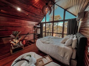 L’intérieur d’un chalet en bois, avec un lit, un luminaire suspendu et une vue sur les arbres à travers de grandes fenêtres.