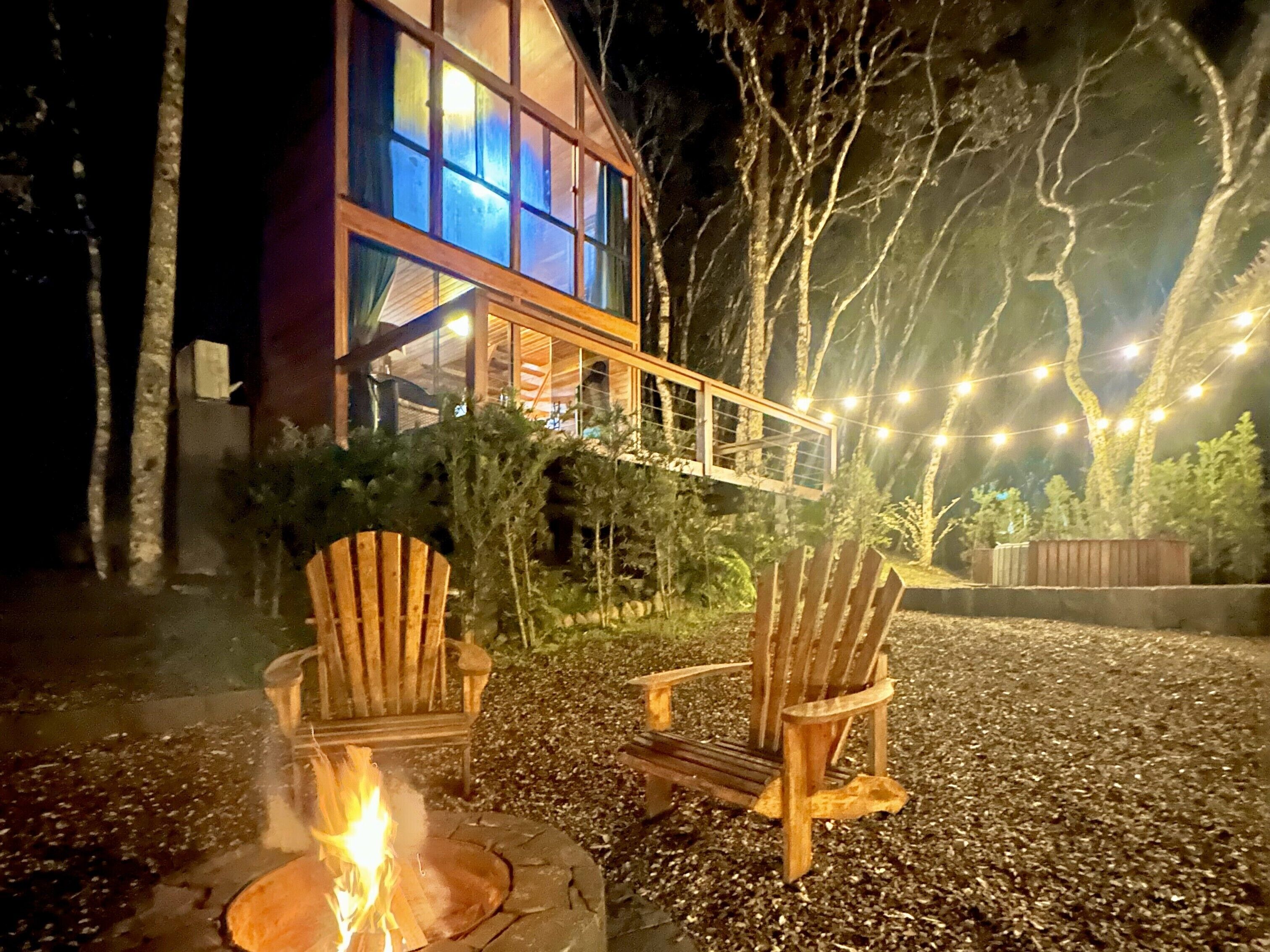 A wooden deck with two Adirondack chairs and a fire pit, surrounded by trees and a building with large windows.