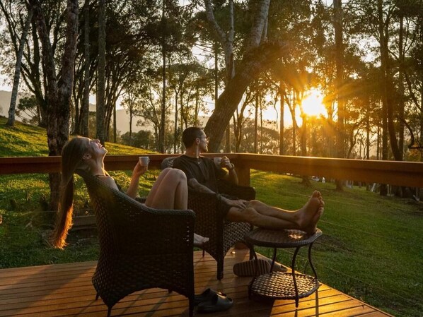 Dos personas descansando en muebles de exterior con vista a árboles y al sol poniéndose.
