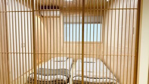 Quarto com divisória de madeira deslizante, duas camas com roupa de cama listrada, uma janela pequena e uma luminária no teto.