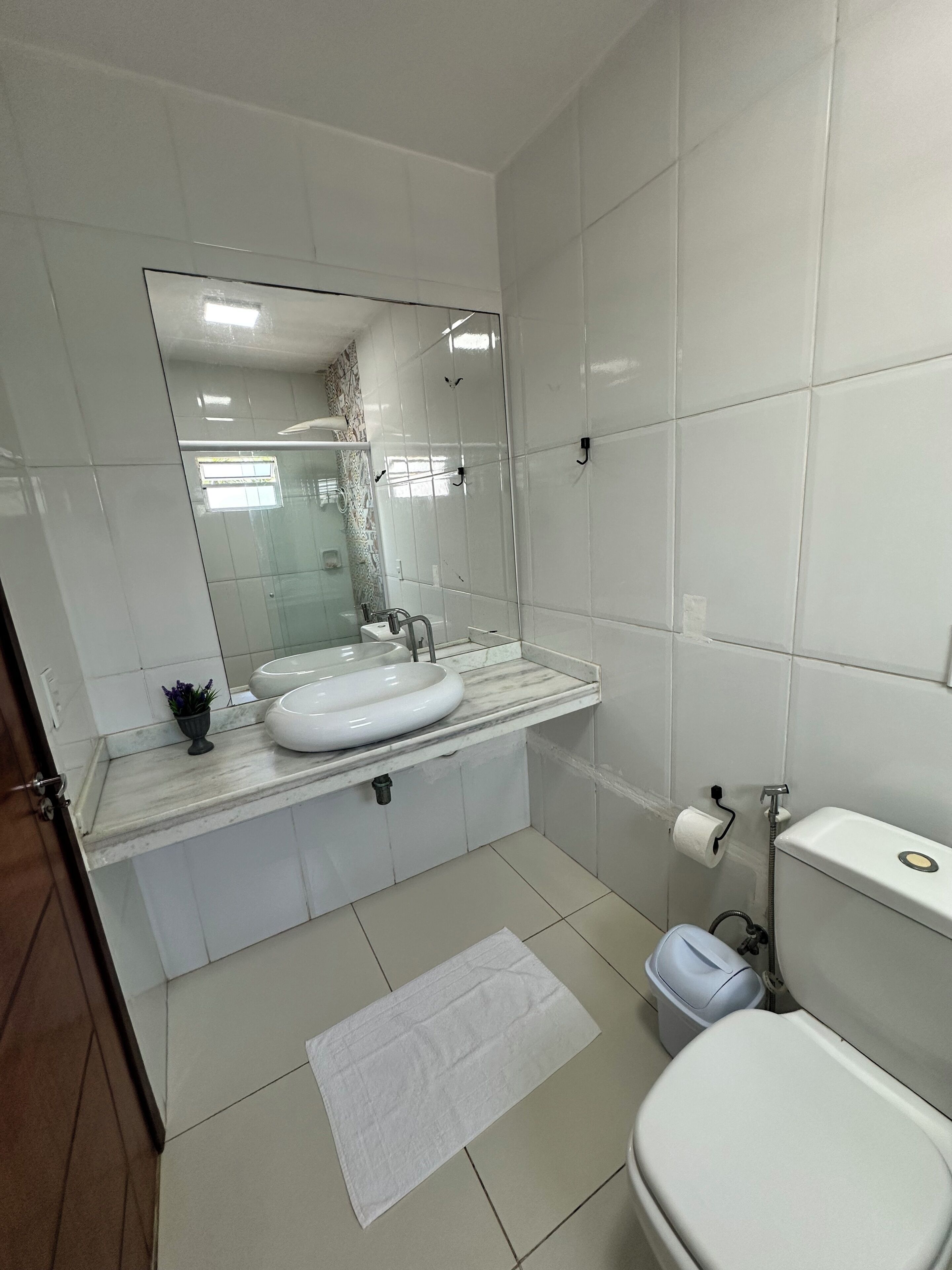 Et badeværelse med hvid vask, spejl, toilet og en lille skraldespand.