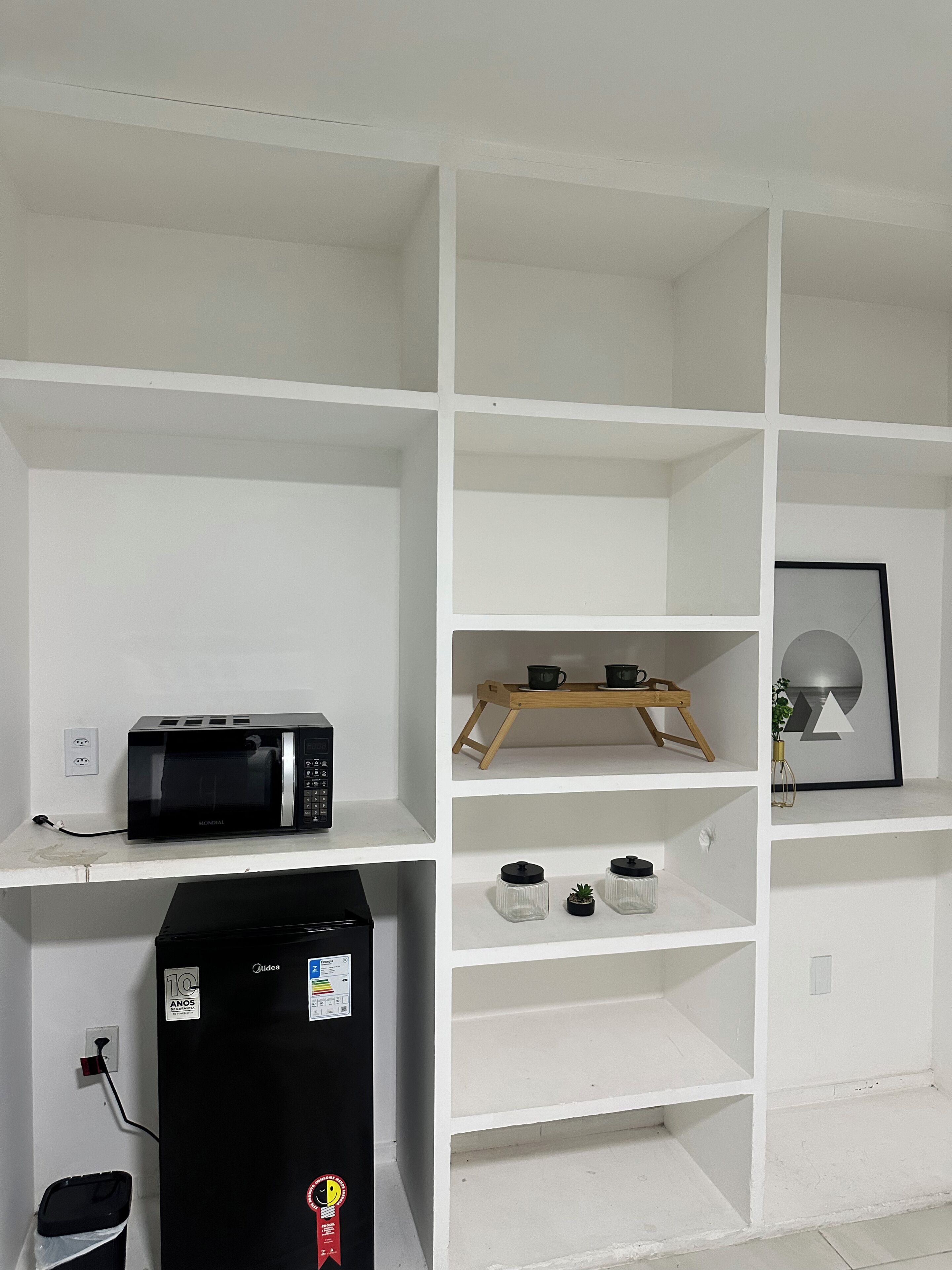 Een witte kamer met links een zwarte magnetron, daaronder een kleine koelkast en rechts een houten plank met een fotolijstje.