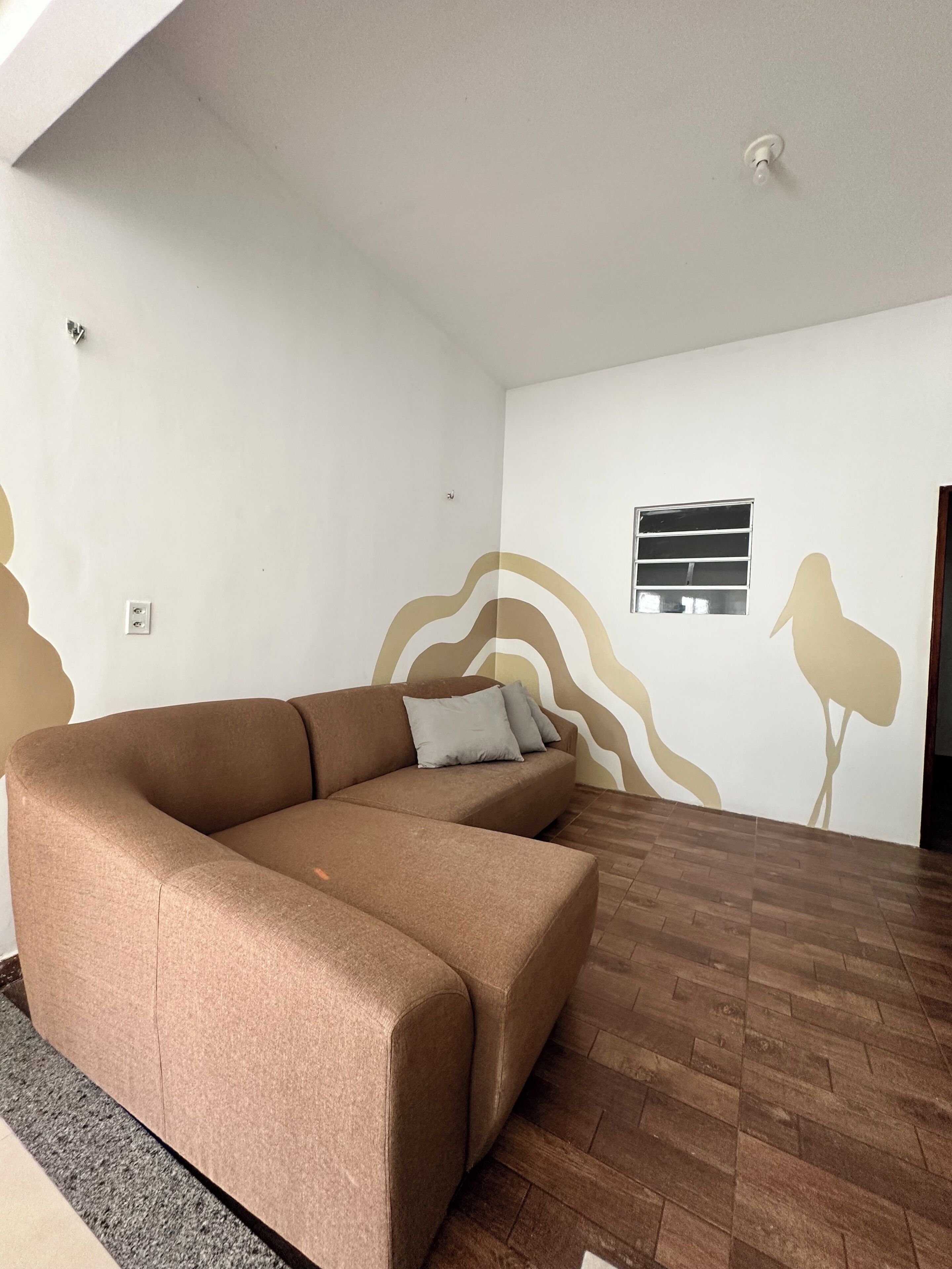 A modern living room with a brown sectional sofa, a small window with blinds, and a decorative wall with abstract patterns.